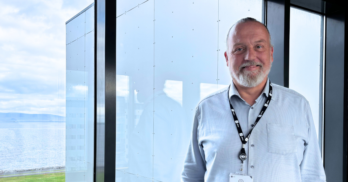 Photo of a man standing with a grey beard and pony tail in a blue shirt in an office.