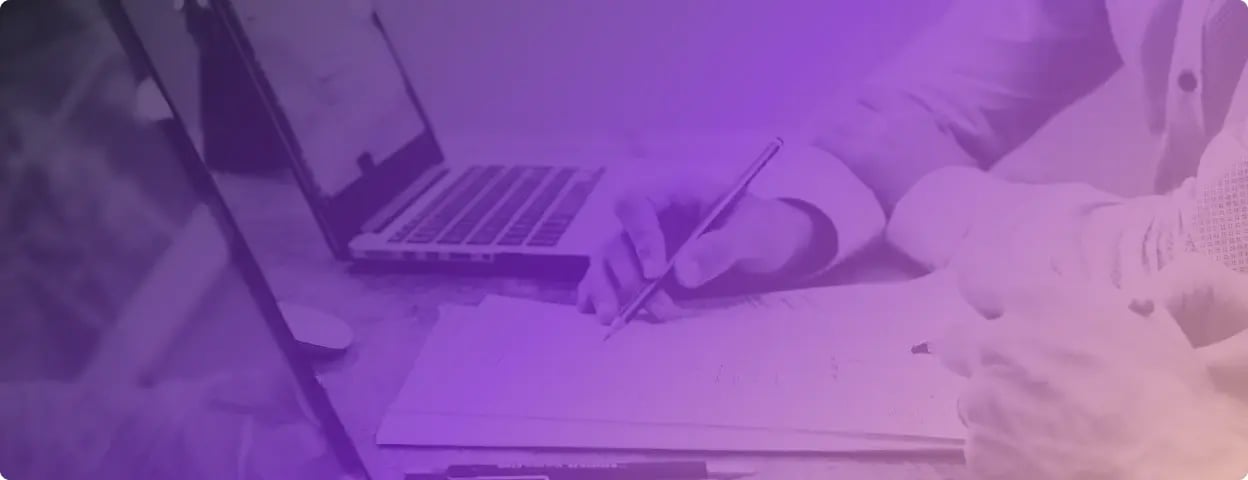 Photo of a laptop on a desk and someone writing on paper